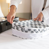 A person closes a Bumkins Packing Cube 2 Pack: Charcoal Check on a bed next to an open suitcase, ideal for travel organization.