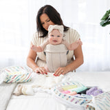 A woman wearing a smiling baby in a carrier folds clothes on a bed to pack into Bumkins Packing Cube 2 Pack: Mickey & Friends Varsity.