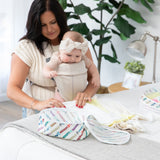 A woman wearing a baby in a carrier folds clothes on a bed to put in Bumkins Packing Cube 2 Pack: Mickey & Friends Varsity.
