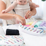 A person with a baby in a carrier organizes clothes in Bumkins Packing Cube 2 Pack: Mickey & Friends Varsity on a white bed.