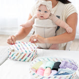 Woman packs baby clothes into Bumkins Disney Packing Cubes & Clear Travel Bags, baby in carrier by her side.