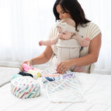 A mom with a baby in a carrier arranges clothes in a Bumkins Packing Cube 2 Pack: Mickey & Friends Varsity on a white bed.