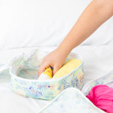 A hand places a rolled yellow shirt into a Bumkins Packing Cube 2 Pack: Aloha Stitch on a white bed.