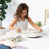 A young girl in a white dress zips Bumkins Packing Cube 2 Pack: Winnie + Friends on a bed.