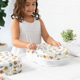 A girl in a white dress smiles, holding a Bumkins Packing Cube 2 Pack: Winnie + Friends, made with waterproof fabric, on the bed.