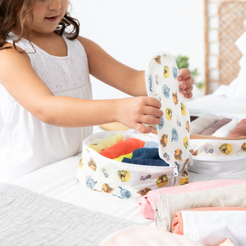 A young girl packs colorful clothes into bags from Bumkins The Outfit Organizer Essential 5-Piece Set: Winnie and Friends on a bed.