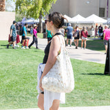 In a park, amid green scenery and people, a person in a black tank top carries a Bumkins Packable Tote Bag: Palm Daze.