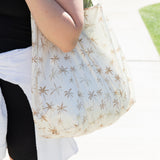A person holds a Bumkins Packable Tote Bag: Palm Daze on a sunny sidewalk.