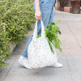 A person in jeans and white sneakers carries a Bumkins Packable Tote Bag: Palm Daze filled with greens, walking on a plant-lined sidewalk.