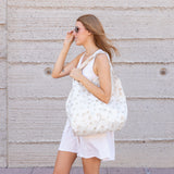 A woman in a white dress and sunglasses walks by a textured wall, carrying Bumkins Packable Tote Bag: Palm Daze.