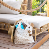 A Bumkins Packable Tote Bag: Palm Daze filled with sunglasses, books, and a straw hat sits by a wicker outdoor sofa on a wooden deck.