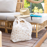 A woven straw hat and books on a wicker sofa sit near Bumkins Packable Tote Bag: Palm Daze on a wooden deck and greenery in the background.