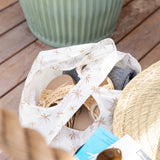 A Bumkins Packable Tote Bag: Palm Daze with flip-flops, hat, and clothes rests on a wooden surface near a green planter.