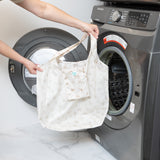 A person holds Bumkins Packable Tote Bag: Palm Daze near an open front-loading washer.