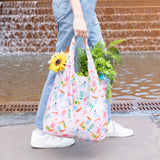 A person in jeans and white sneakers carries Bumkins Packable Tote Bag: Boots filled with flowers and vegetables, strolling past a brick wall.