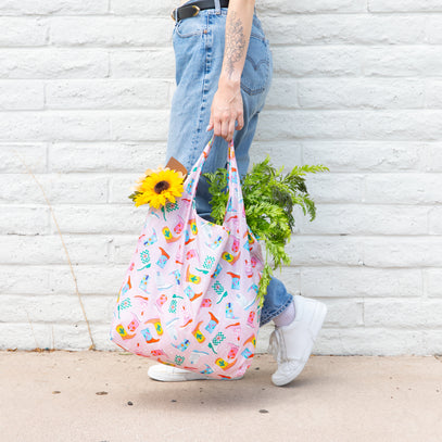 A person walks against a white brick wall, carrying a colorful tote bag filled with a sunflower and green foliage. They wear blue jeans, a black belt, white sneakers, and have a visible arm tattoo.