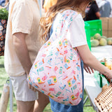 A person with a Bumkins Packable Tote Bag: Boots browses fresh fruits at an outdoor market.