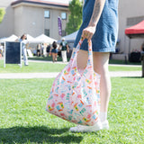 A person carries a vibrant Bumkins Packable Tote Bag: Boots in a lively park, with tents and people in the background.