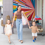 A woman walks with two kids, holding a Bumkins Packable Tote Bag: Boots, near a colorful mural. Its sunny and they are all smiling.
