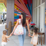 A woman with a Bumkins Packable Tote Bag: Boots and two children strolls by a mural near a cactus-shaped art piece.