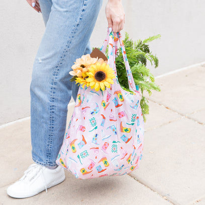 A person walks on a pavement carrying a colorful reusable shopping bag adorned with various small patterns. The bag contains groceries, including a sunflower, some greens, and a baguette. The person is wearing blue jeans and white sneakers.