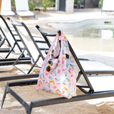 A Bumkins Packable Tote Bag: Boots hangs on a pool lounge chair. Sunglasses dangle from its side, with empty chairs behind.