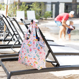 A Bumkins Packable Tote Bag: Boots hangs on a lounge chair by the pool while someone holds a child near the water.