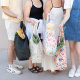 Four people hold bags filled with groceries against a light background. One holds Bumkins Packable Tote Bag: Boots filled with popcorn.