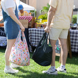 Two people holding bags, one a Bumkins Packable Tote Bag: Boots, at an outdoor market, with a stall and products displayed behind them.