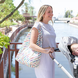 A woman in a light gray outfit with a Bumkins Packable Tote Bag: Mickey & Friends Varsity on her shoulder pushes a stroller by a canal.