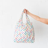 A hand holds a Bumkins Packable Tote Bag: Mickey & Friends Varsity against a white brick wall.