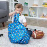 A young boy sits with his Bumkins Packable Tote Bag: Mickey & Friends Forever, next to a football against toy-filled shelves.