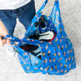 A child stands on a light carpet with a Bumkins Packable Tote Bag: Mickey & Friends Forever holding shoes and sports gear.