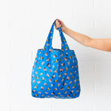 A hand holds a Bumkins Packable Tote Bag: Mickey & Friends Forever against a white brick wall.