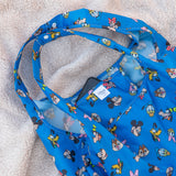 Close-up of Bumkins Packable Tote Bag: Mickey & Friends Forever on a beige blanket with a smartphone in the pocket.