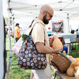 A man with a Bumkins Packable Tote Bag: Ghostly Gallery over his shoulder examines a butternut squash at an outdoor market under a white tent.