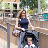 A woman smiles as she carries a Bumkins Packable Tote Bag: Ghostly Gallery and pushes a stroller outdoors, surrounded by trees and railings.