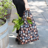 A person carries a Bumkins Packable Tote Bag: Ghostly Gallery filled with groceries and carrots, walking on a paved path.
