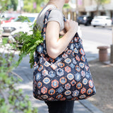 A person carries a Bumkins Packable Tote Bag: Ghostly Gallery, filled with green veggies on a bustling city street.