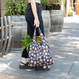 A person in black jeans holds a Bumkins Packable Tote Bag: Ghostly Gallery filled with veggies, walking past wooden barrels on a paved path.