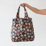 A hand holds a Bumkins Packable Tote Bag: Ghostly Gallery against a white brick wall.