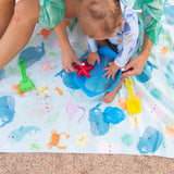 A child plays on a Bumkins Reversible Roll & Go Blanket, Rolling with the Waves on the sand with toy shovels and a starfish mold.