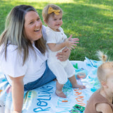 A woman and toddler sit on a Bumkins Reversible Roll & Go Blanket, Rolling with the Waves on the grass outdoors.