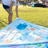 A person lays a Bumkins Reversible Roll & Go Blanket, Rolling with the Waves on the grass at an outdoor event.