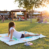 A girl relaxes on a Bumkins Reversible Roll & Go Blanket, Rolling with the Waves in a park at sunset, amidst people and food trucks.