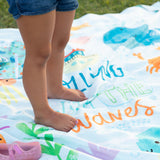 A child stands barefoot on a Bumkins Reversible Roll & Go Blanket, Rolling with the Waves, next to pink sandals on grass.