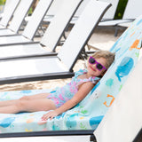 A young girl in sunglasses and swimsuit relaxes on a Bumkins Reversible Roll & Go Blanket, Rolling with the Waves among empty beach chairs.