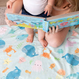 A child sits on an adult's lap, reading a book on a Bumkins Reversible Roll & Go Blanket, Rolling with the Waves.