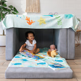 A child smiles brightly in a gray cushion fort with Bumkins Reversible Roll & Go Blanket, Rolling with the Waves, plush toys, and books.