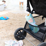 Close-up of a stroller on a sandy beach with a rolled Bumkins Reversible Roll & Go Blanket, Rolling with the Waves in the basket.